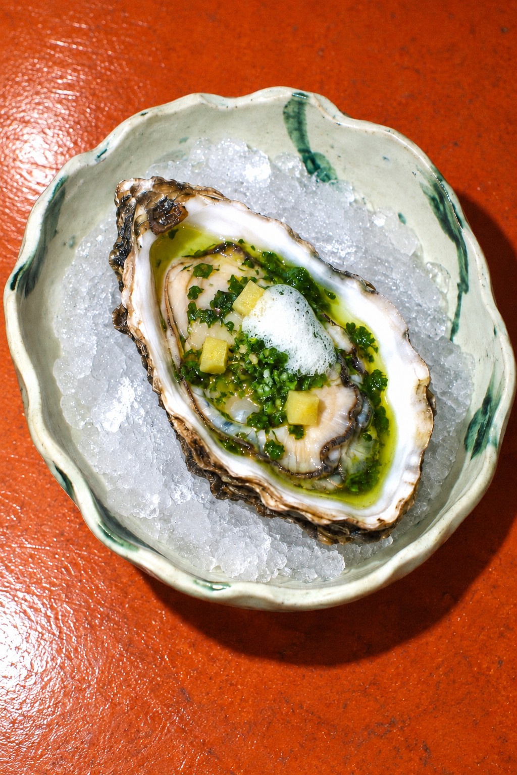 One oyster with green chermoula, preserved lemon and lemon foam on crushed ice in celadon porcelain on an orange table.