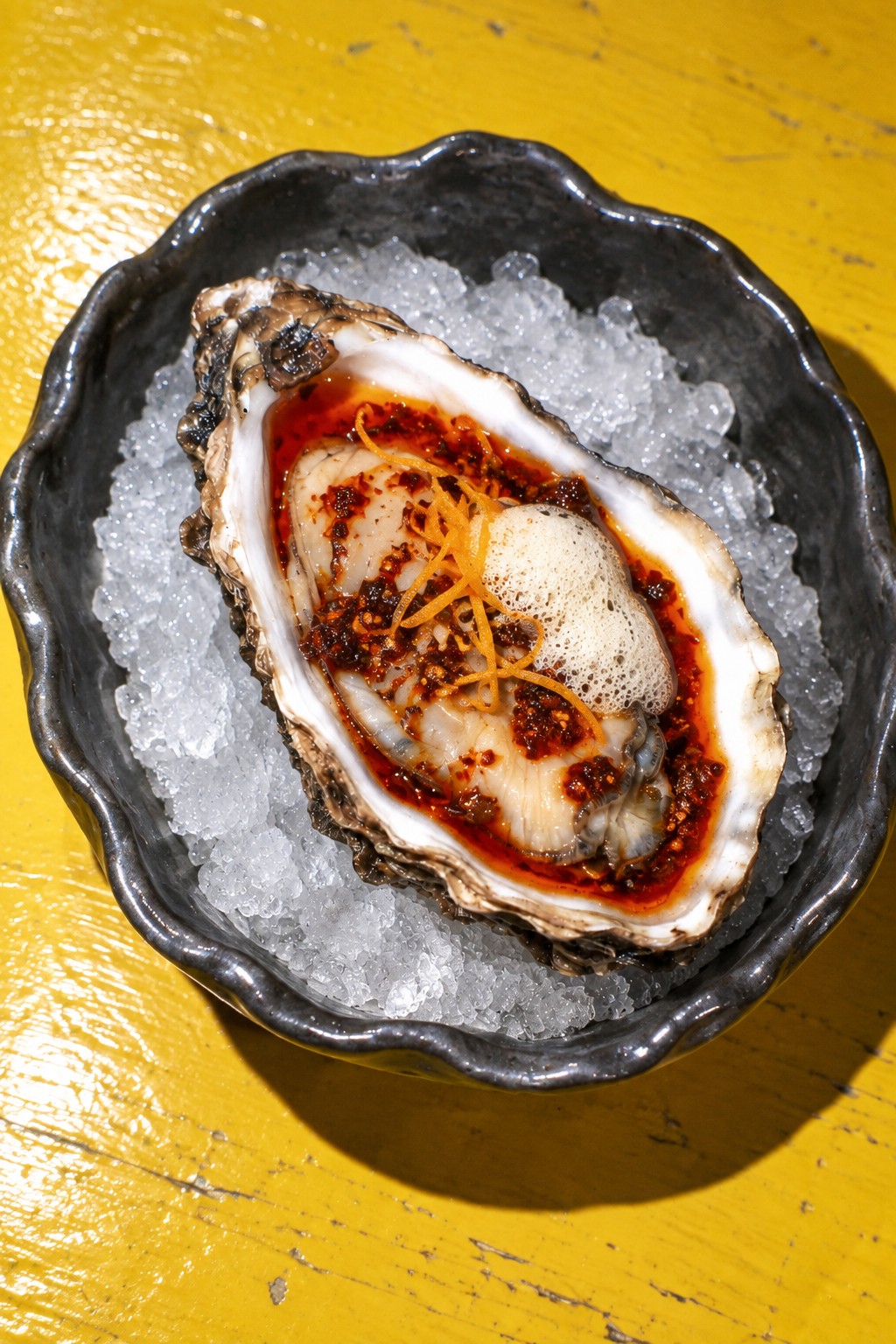 One oyster in ruby five-spice chilli oil with ginger foam and orange zest, on crushed ice in charcoal porcelain on a yellow table.