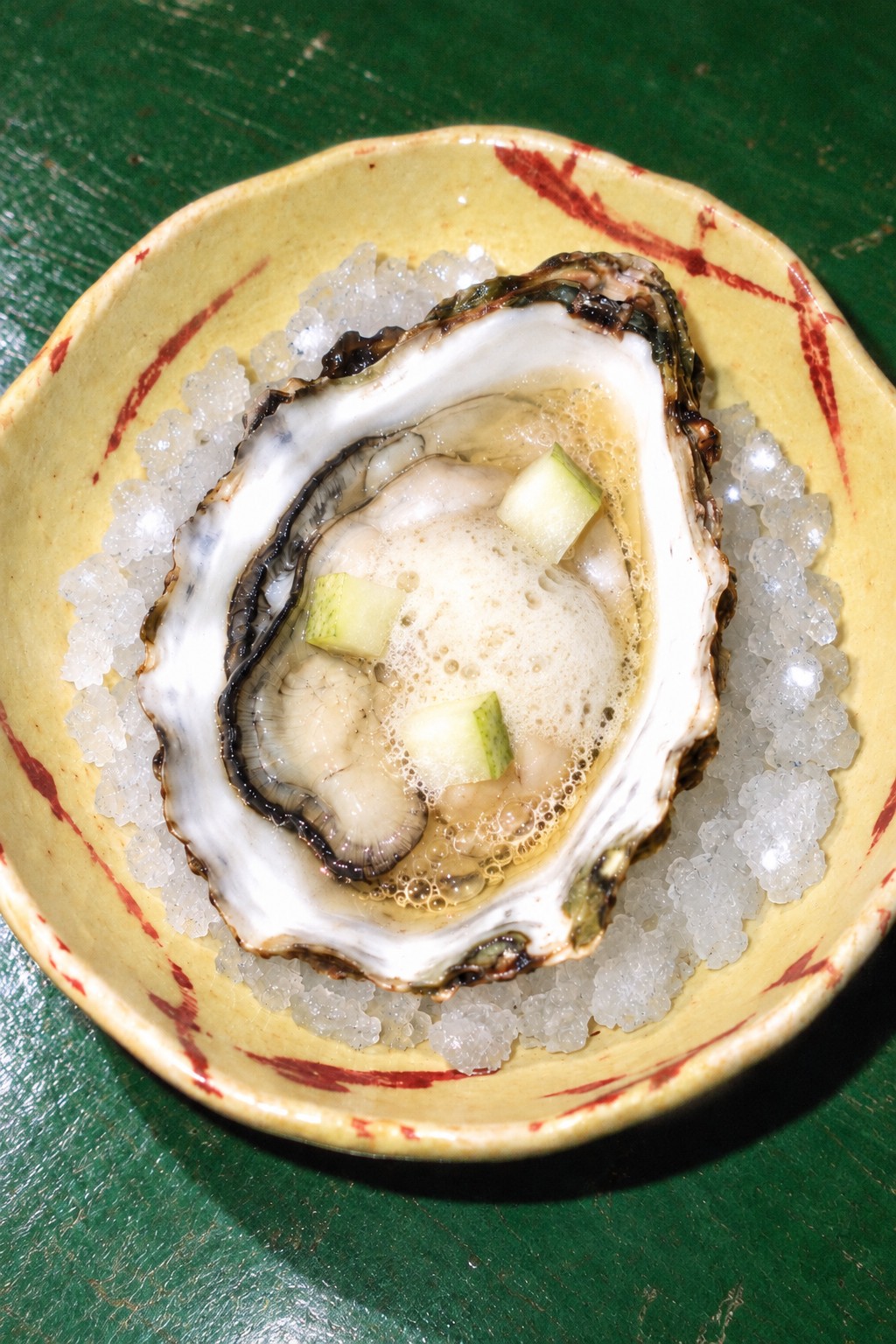 One oyster with pear foam, fresh pear and poiré on crushed ice in yellow porcelain on a green table, without herbs.