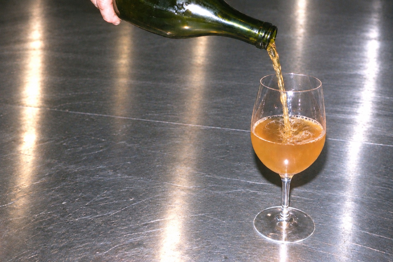 A green bottle pouring orange wine into a glass on a stainless-steel counter.