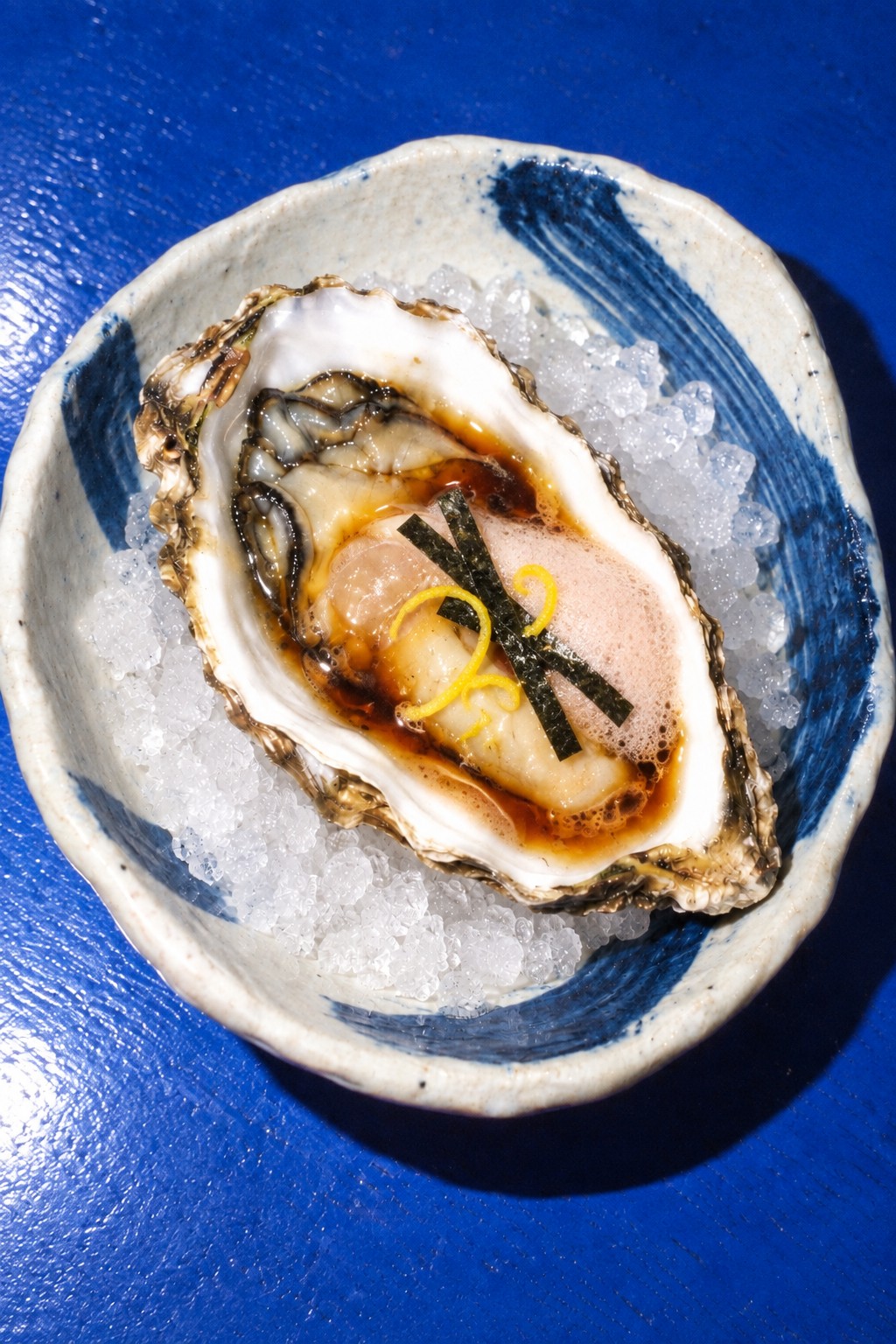 One oyster with dark shiitake reduction, miso, pale plum foam, nori and yuzu zest on crushed ice in blue-brushed Japanese porcelain on a cobalt table.