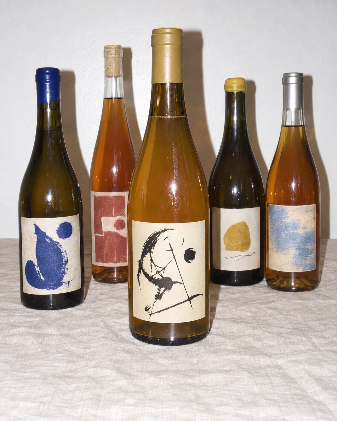 Five orange-wine bottles with different illustrated labels on a linen-covered table against a white wall, photographed with direct flash.