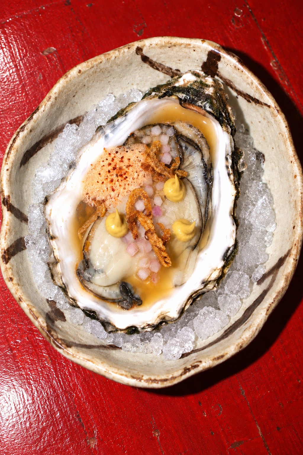 One oyster with chilli foam, natural sweet-mustard dollops and crisp onion, on crushed ice in ivory porcelain on a red table.
